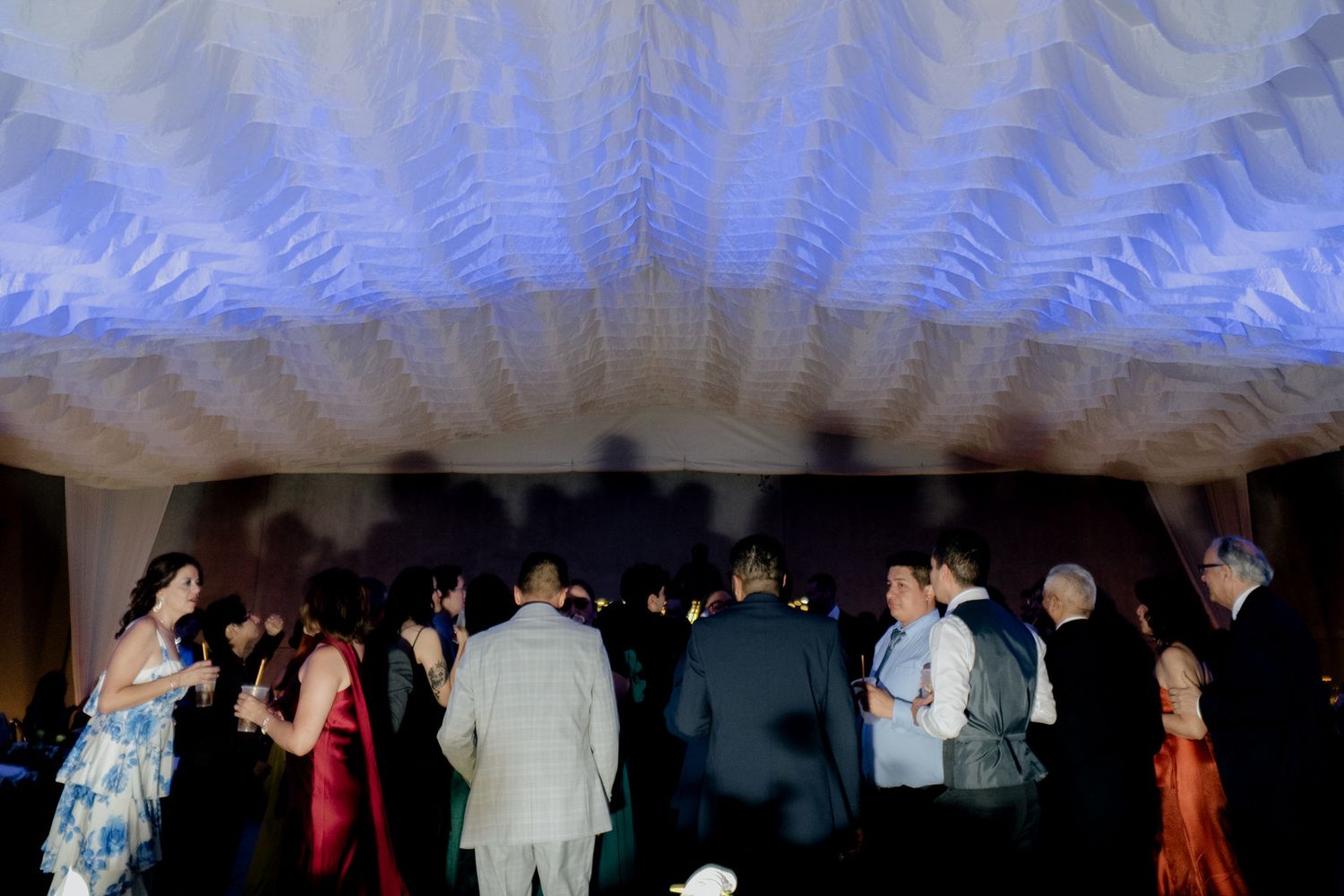 Wedding guests mingle and socialize under blue-lit decorative ceiling at elegant reception venue.