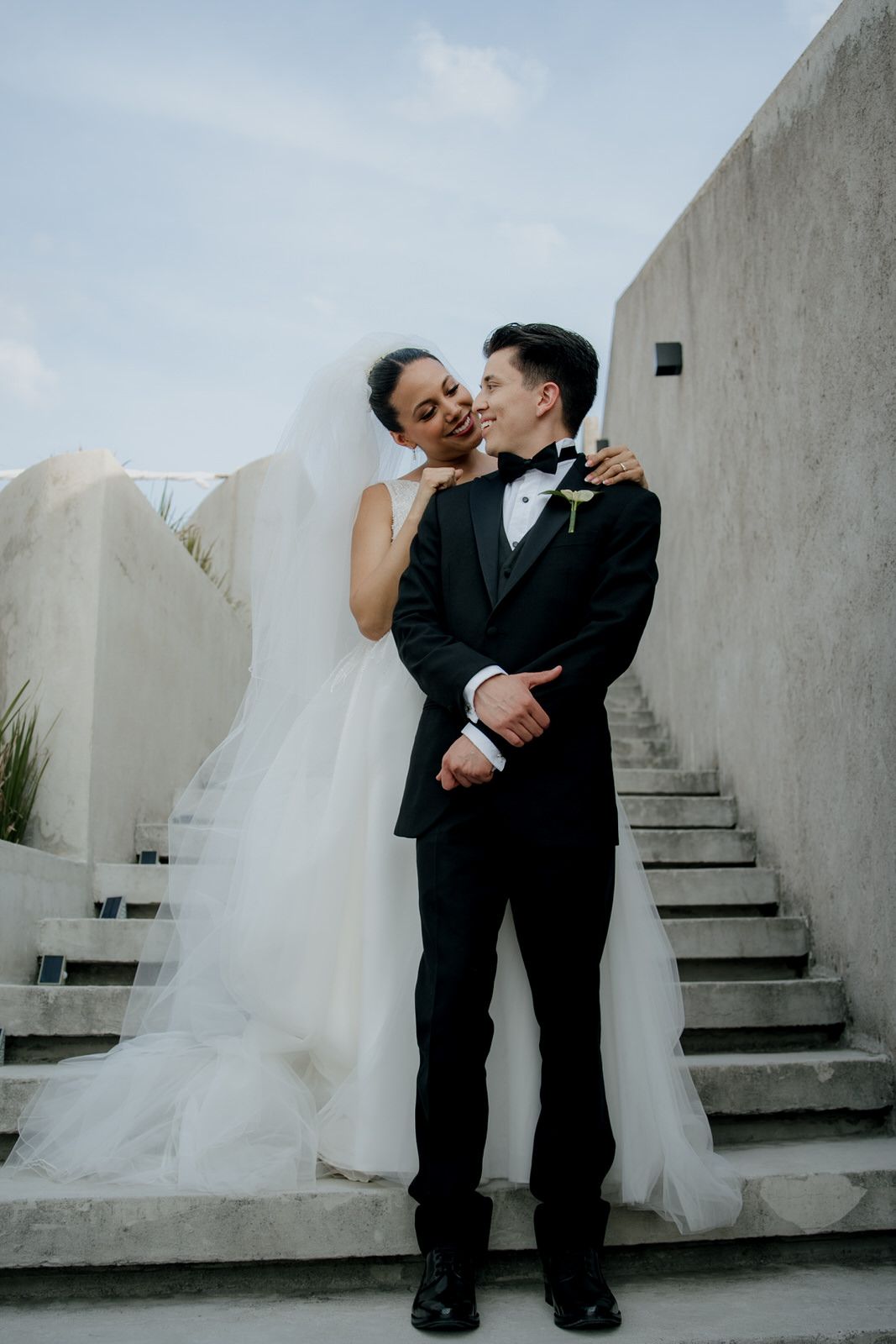 Bride and groom share a romantic kiss on concrete stairs, bride in white gown with veil, groom in black tuxedo.