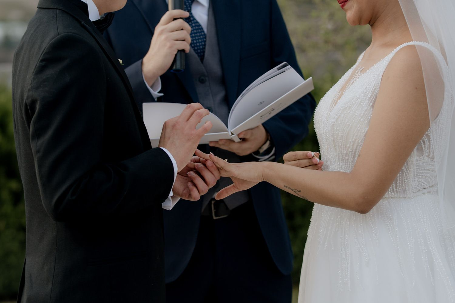 Bride and groom exchanging rings during outdoor wedding ceremony with officiant holding book.
