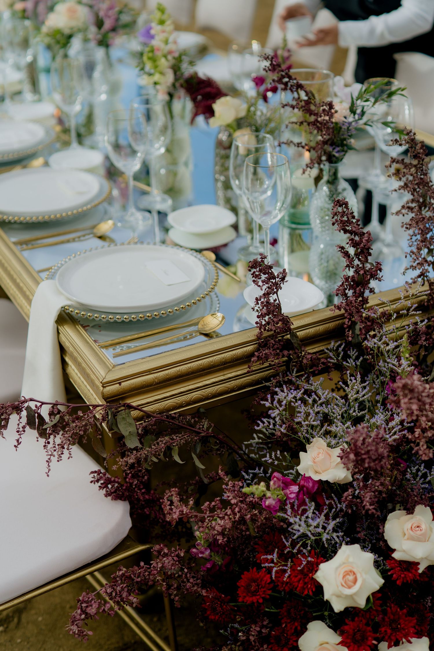 Elegant wedding table setting with gold frames, white plates, crystal glasses, and lush burgundy floral arrangements.
