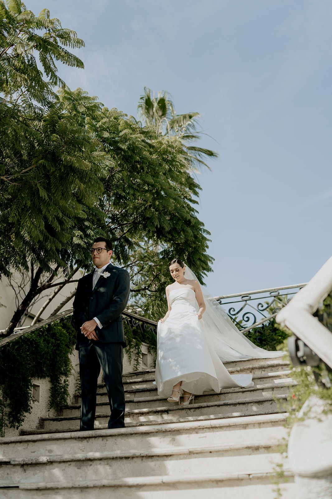 Bride and groom standing on elegant outdoor stone steps surrounded by lush green trees on wedding day.