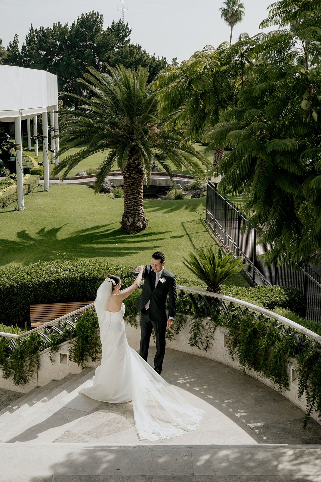 Bride and groom descend elegant curved stairs in a lush garden, groom holding bride's hand gracefully.