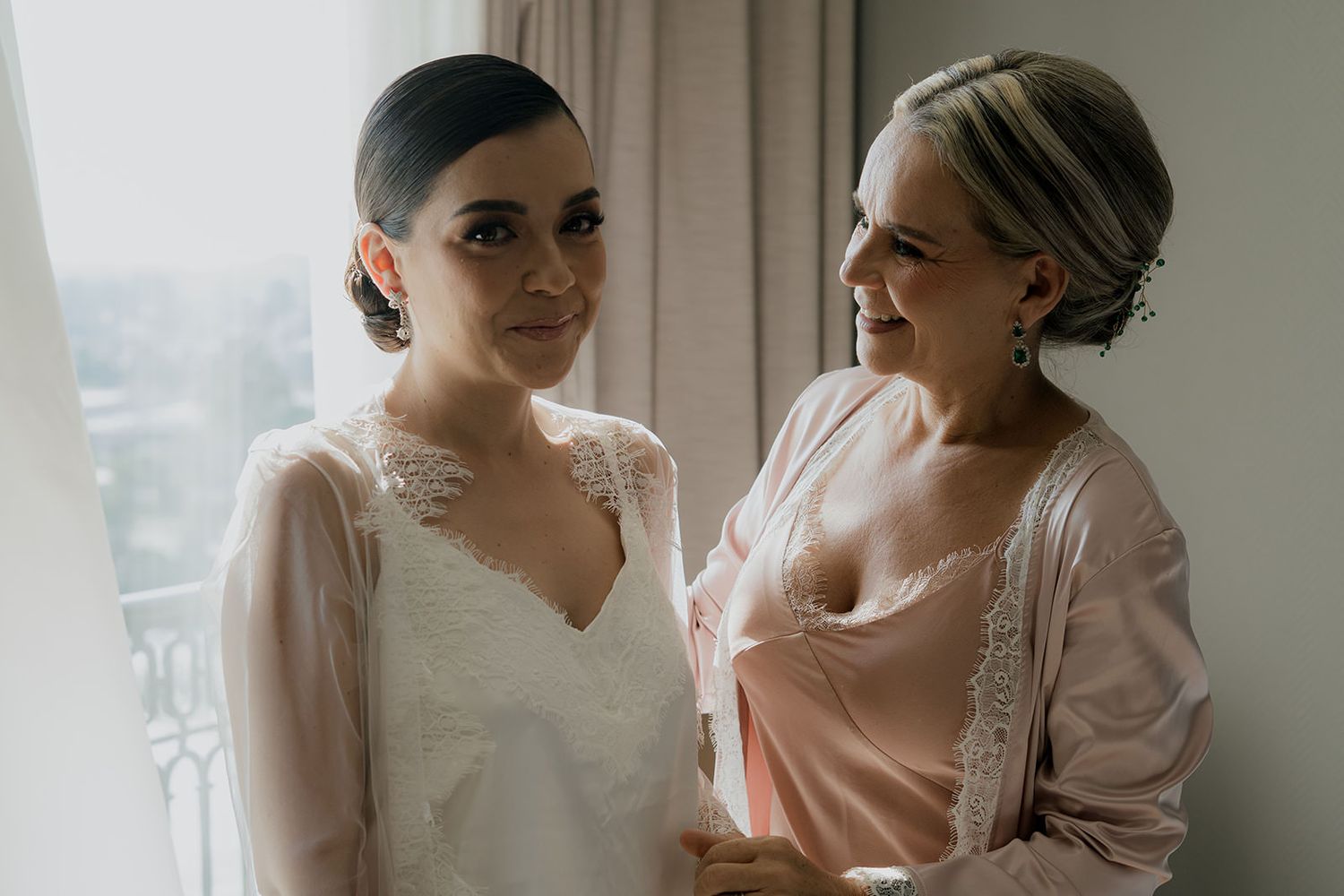 Bride in white lace wedding dress being helped by her mother in a pink robe near a bright window.