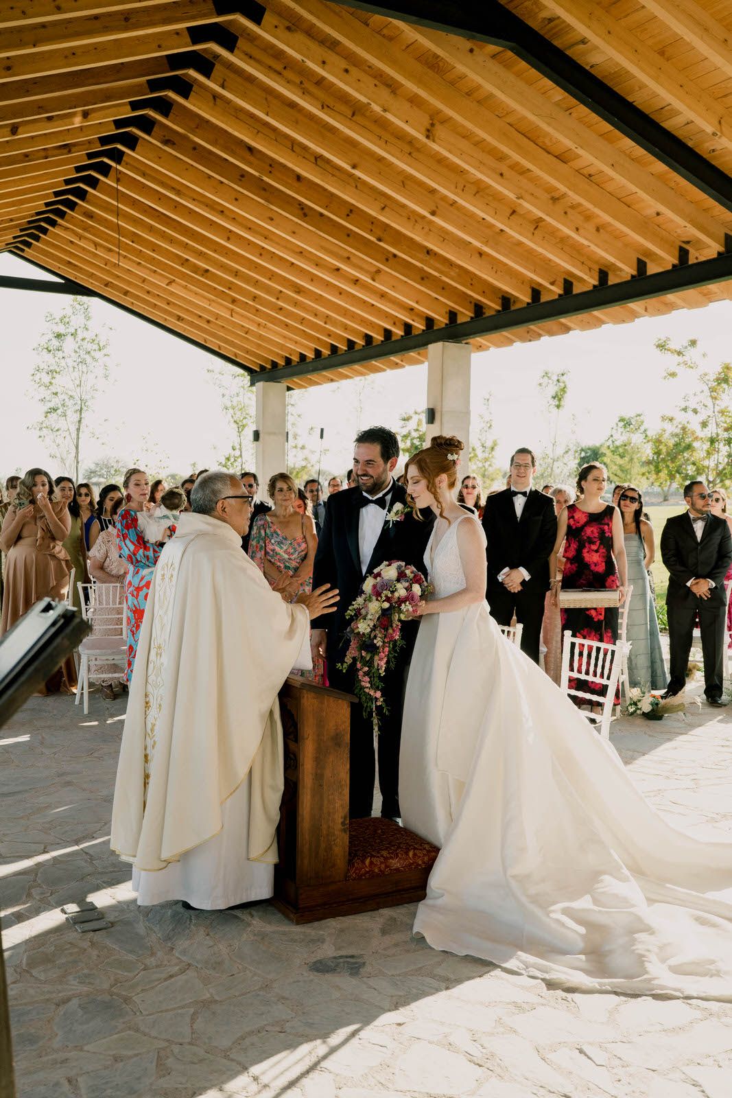 Religious wedding ceremony taking place under wooden pergola with formal dressed guests watching.
