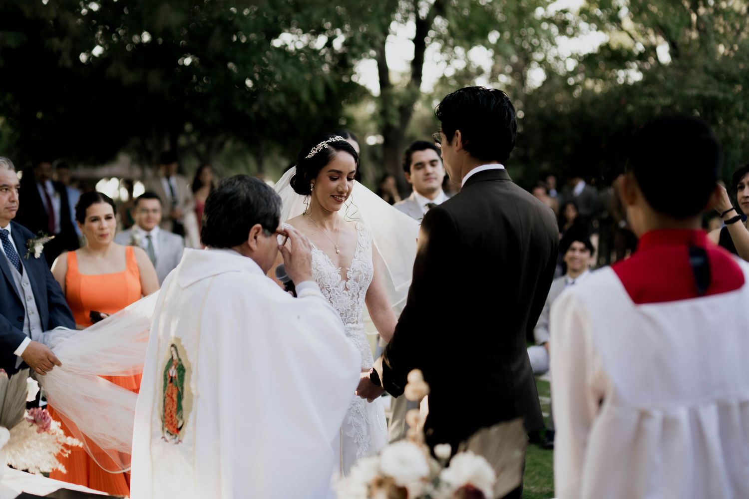 Wedding guests in formal attire gathered at an outdoor venue with natural landscaping.
