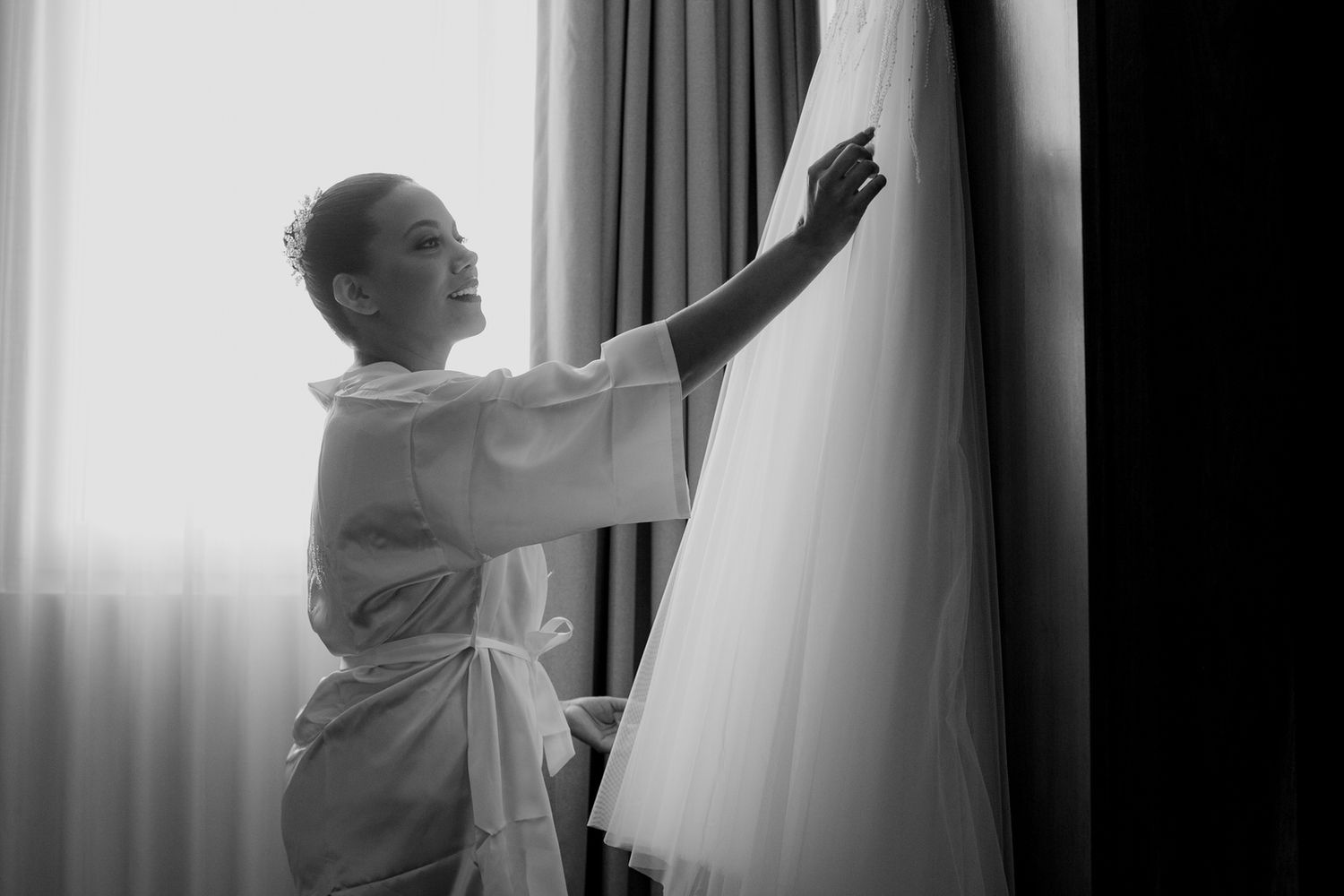 Bride in white robe admiring her wedding dress hanging by a window in a black and white photo.