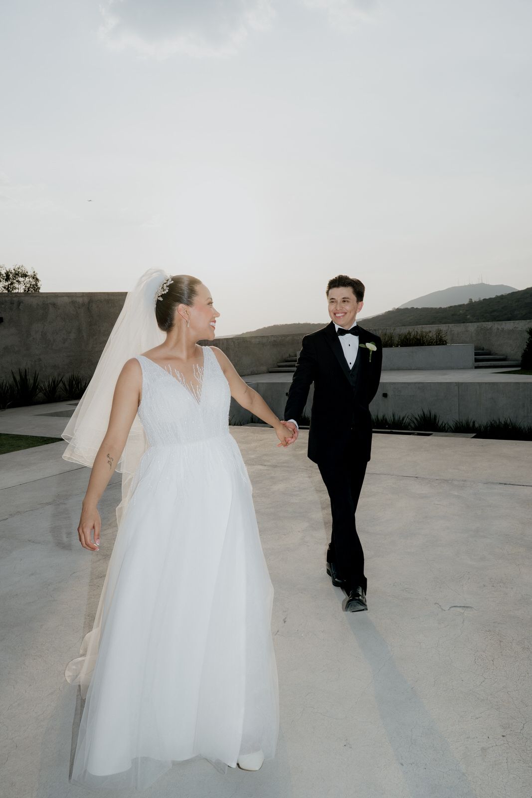 Bride in white tulle gown and veil holds hands with groom in black tuxedo walking at outdoor wedding venue.