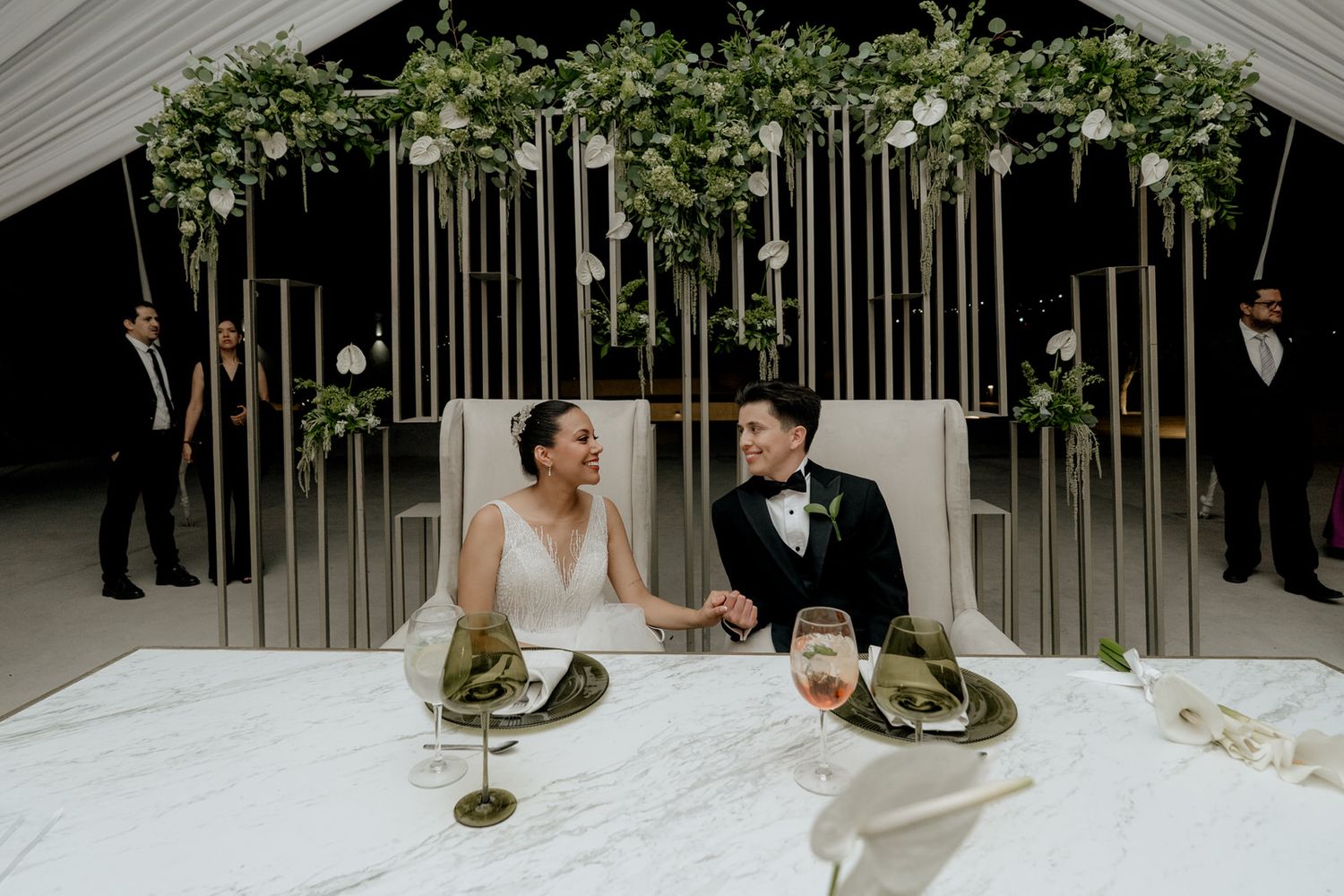 Newlyweds hold hands at elegantly decorated sweetheart table under floral wedding arch.