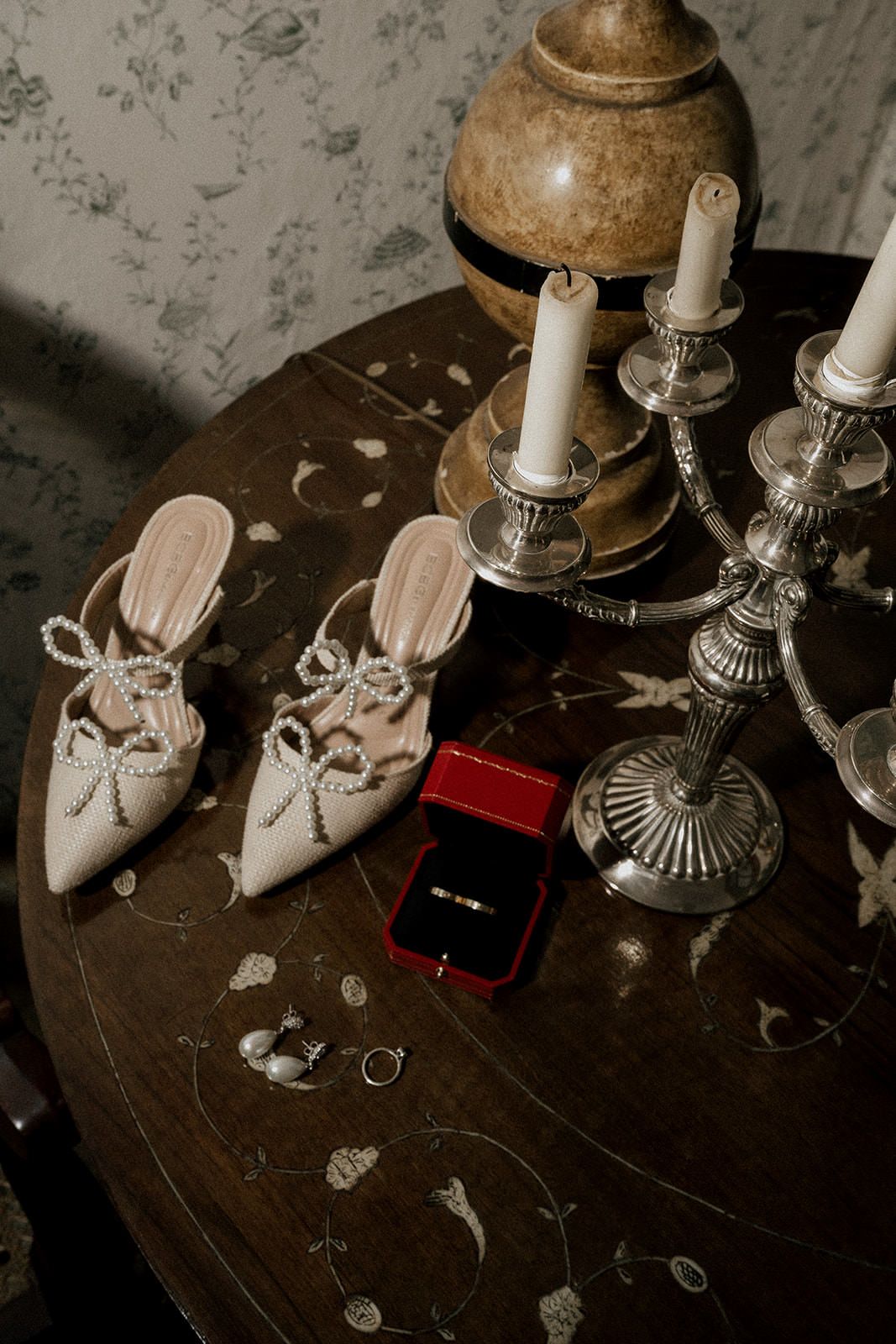 Wedding accessories including bridal shoes and rings arranged with candles on a dark wooden table.
