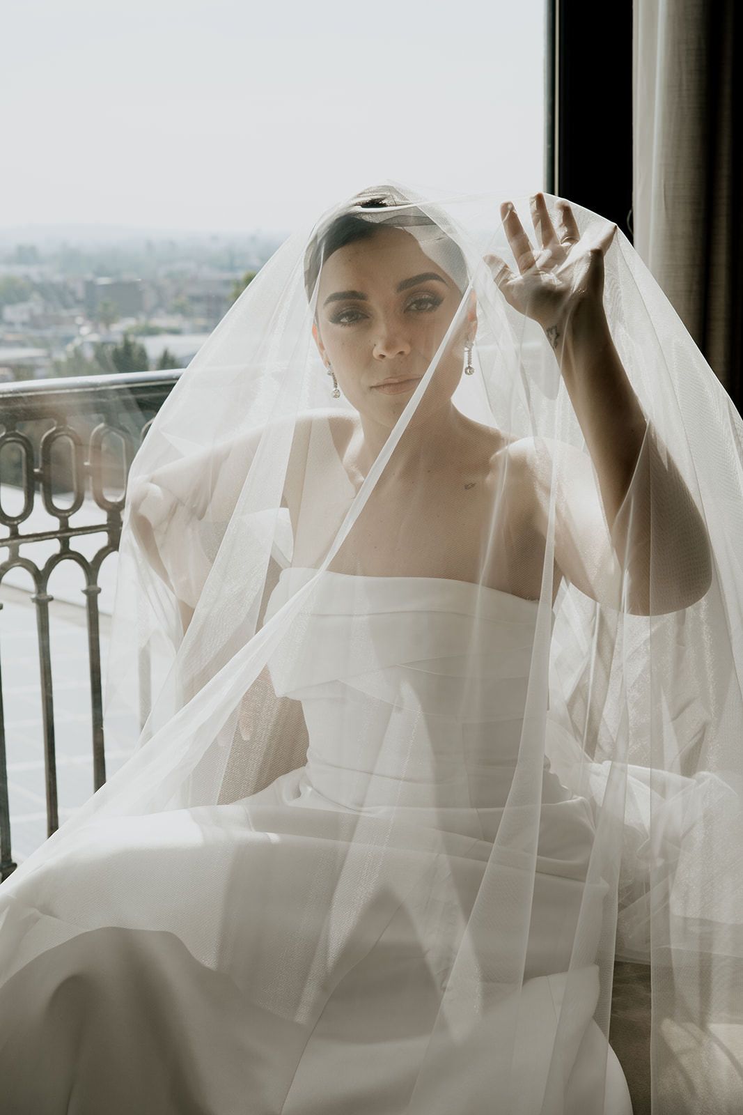 Elegant bride in a white gown and veil sitting on a balcony, gracefully lifting her hand in soft light.