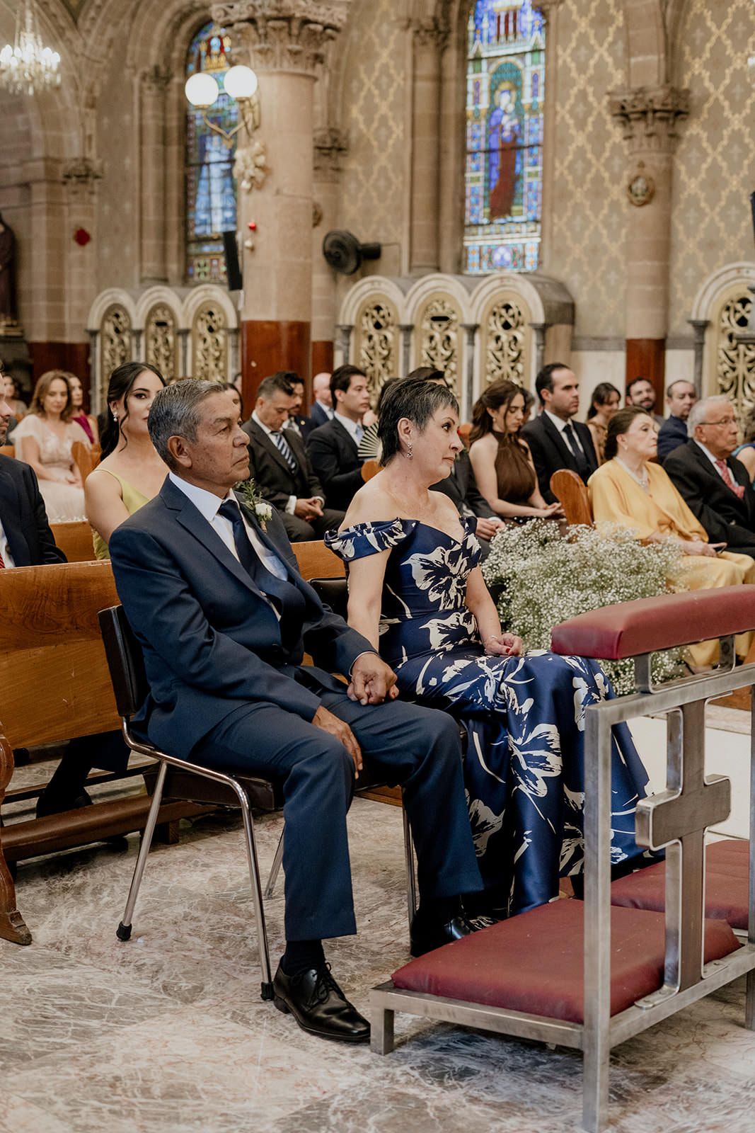 Guests seated in church pews during a wedding ceremony, woman in floral dress beside man in navy suit.