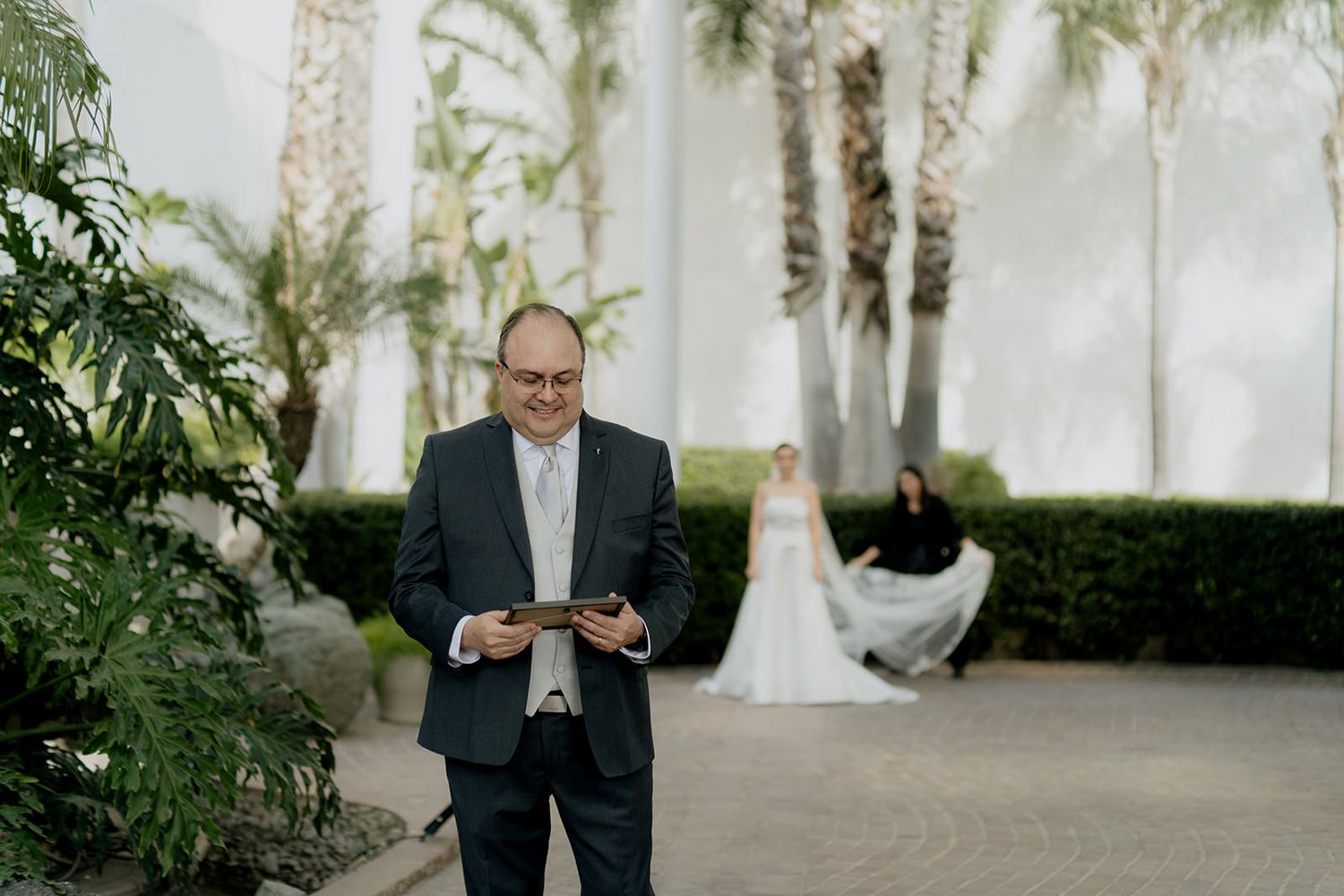 Groom reads vows from a tablet outdoors, with bride in white gown standing among palm trees in background.