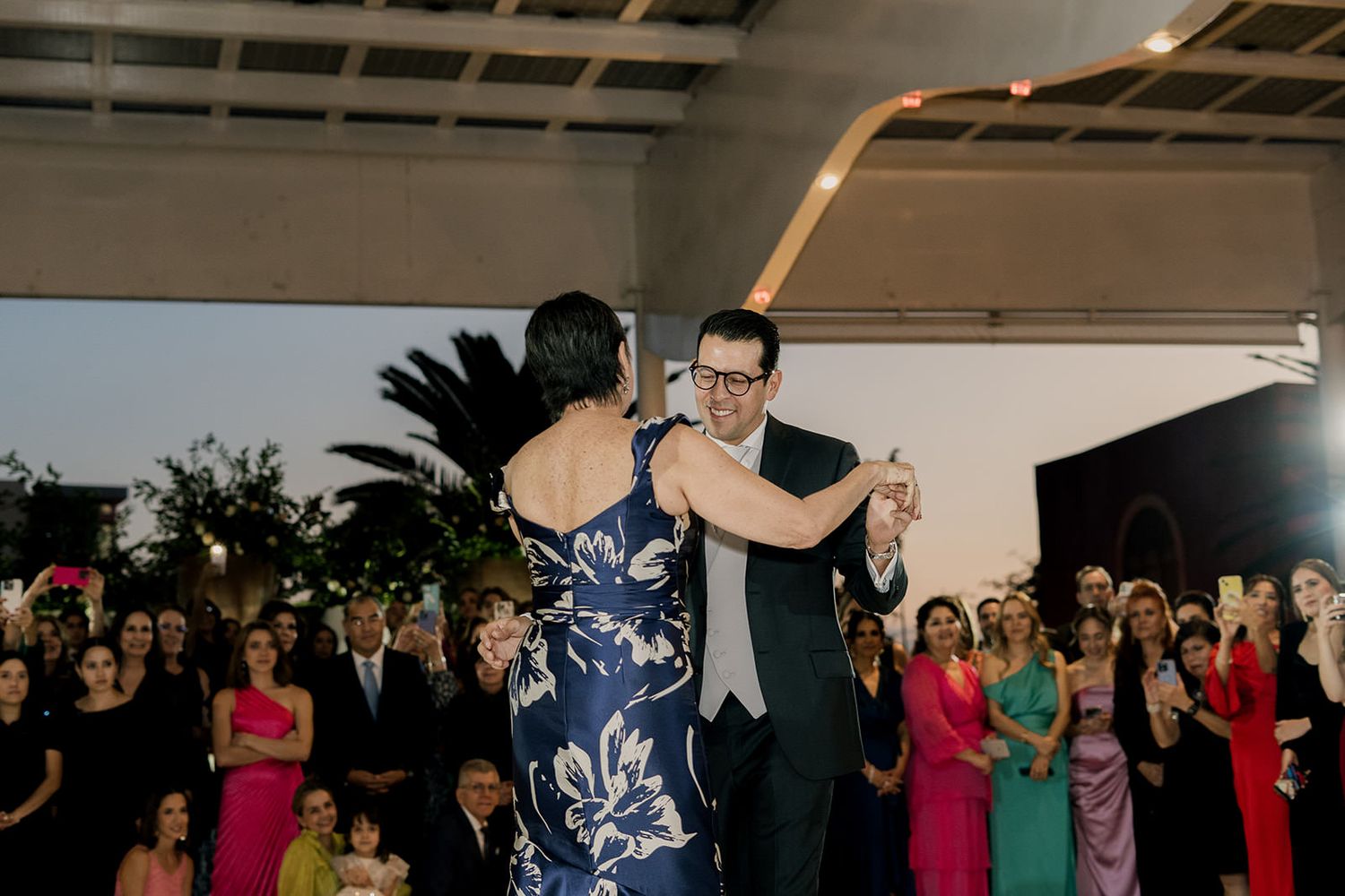 Groom dancing with mother in stunning blue floral gown at elegant outdoor wedding reception at dusk.