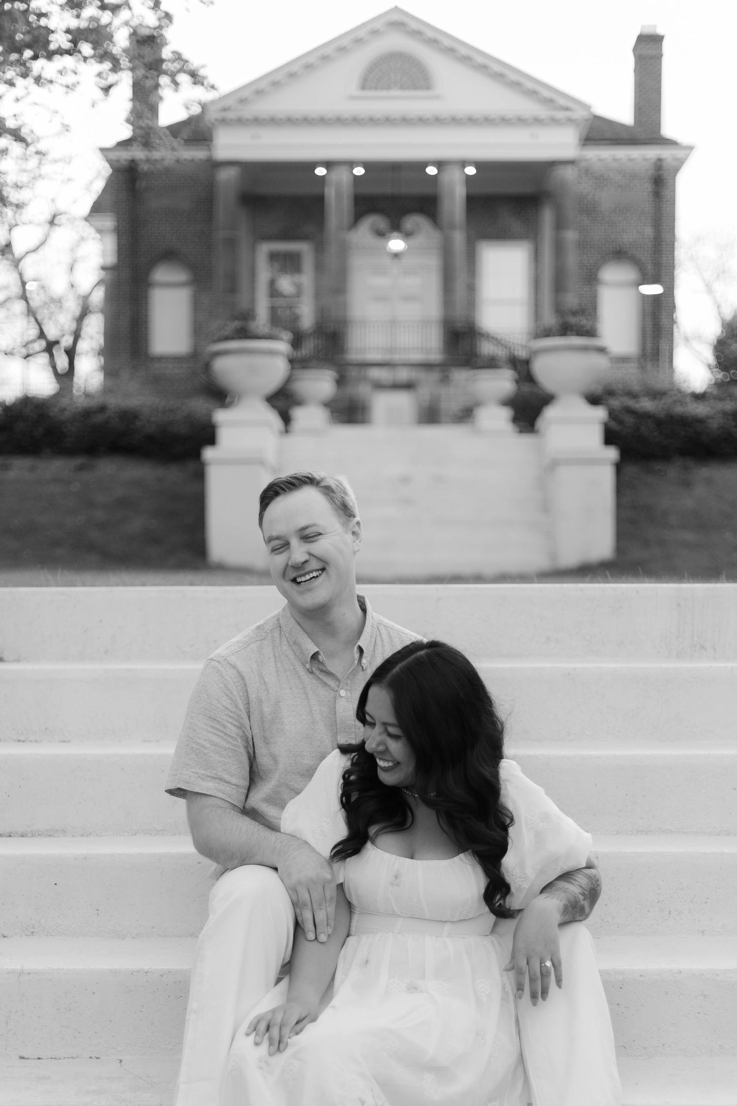 Black and white photo of a smiling couple sitting on steps in front of a classic brick mansion.