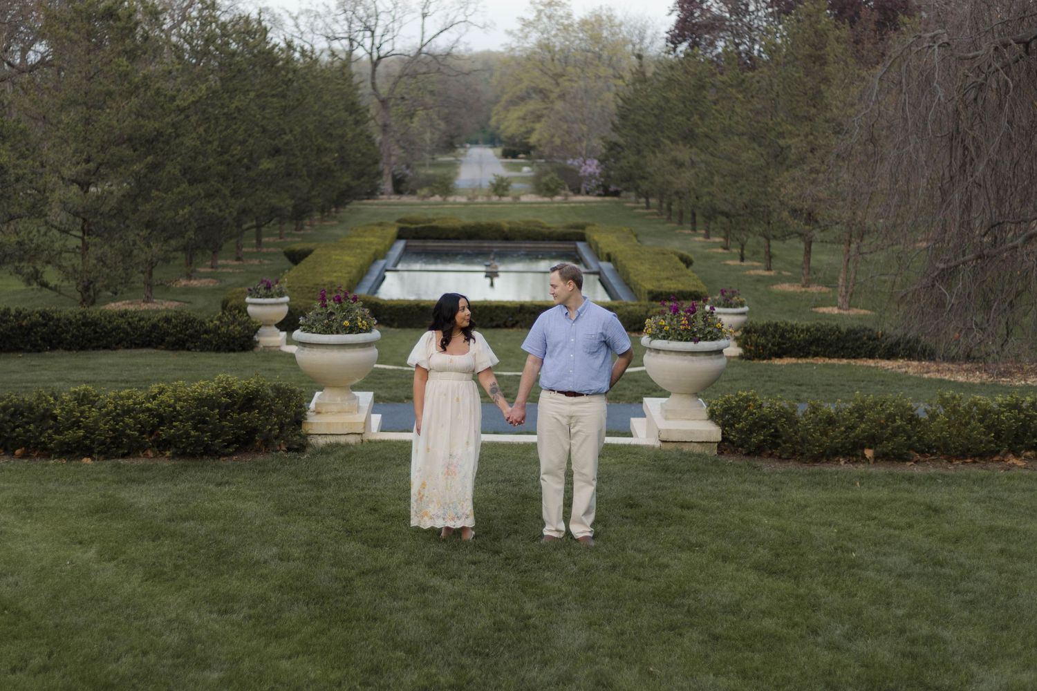 Couple holding hands in formal garden with fountain, lush hedges, and manicured lawn during outdoor maternity photoshoot.
