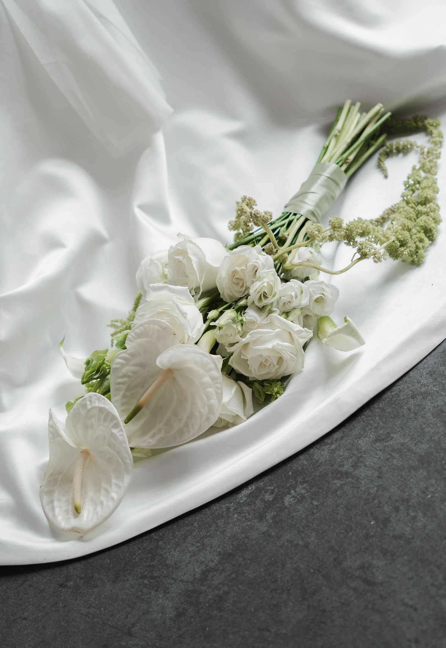 Elegant white wedding bouquet with anthurium, roses, and greenery arranged on white satin fabric against gray surface.
