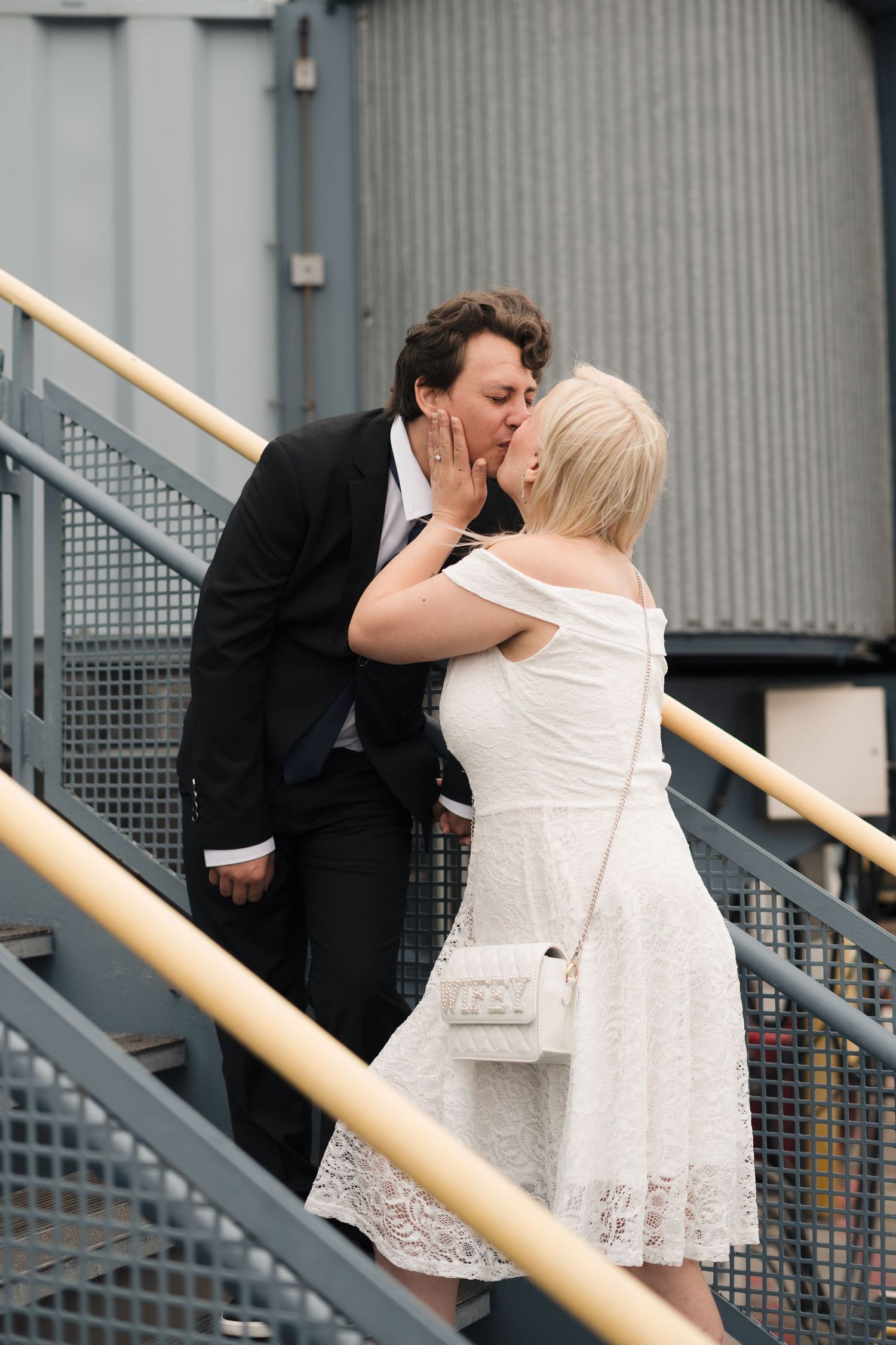 Couple sharing a romantic kiss on metal stairs, groom in black suit and bride in white lace dress with small purse.