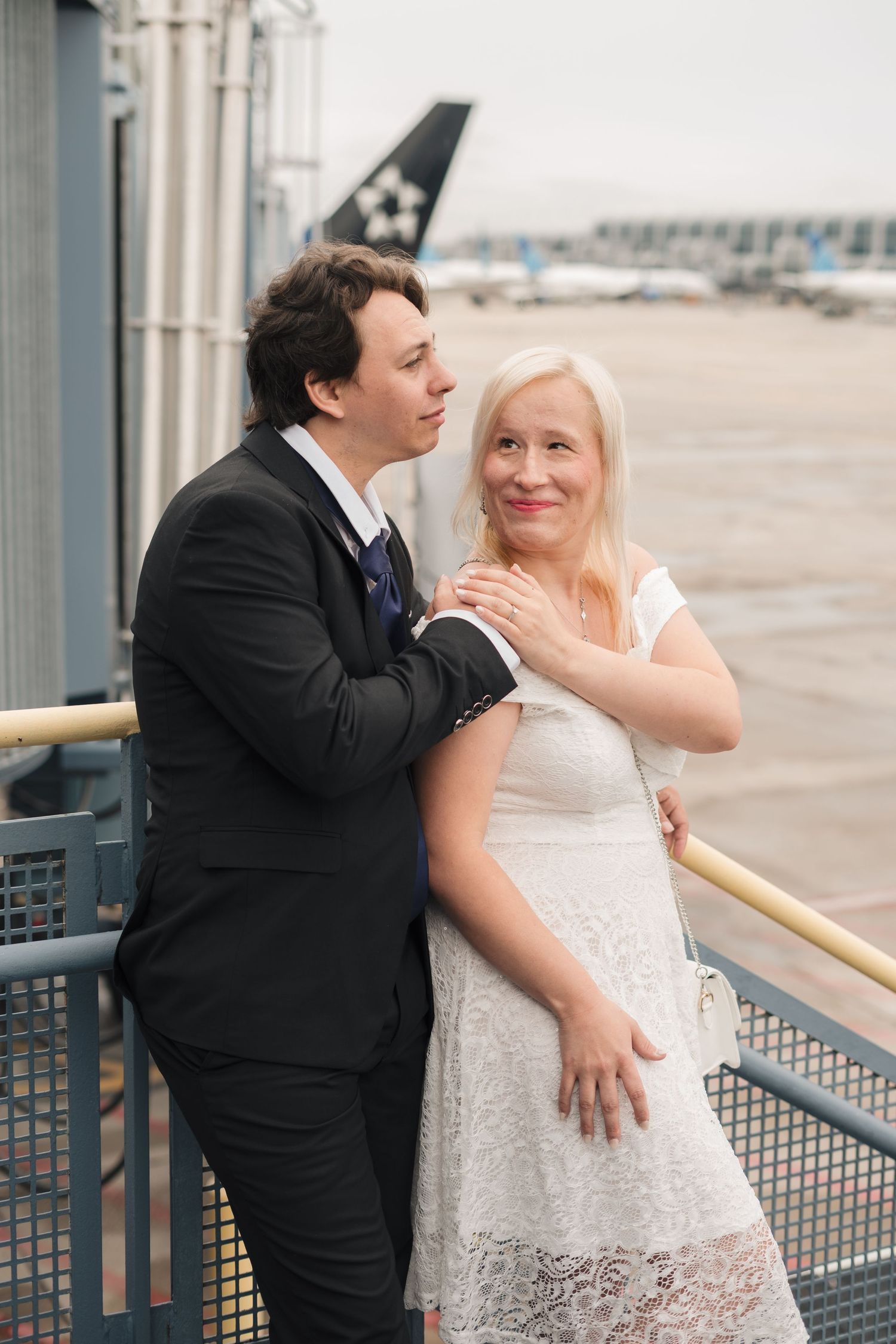 Couple in wedding attire embracing on airport tarmac balcony, man in suit and woman in white lace dress.