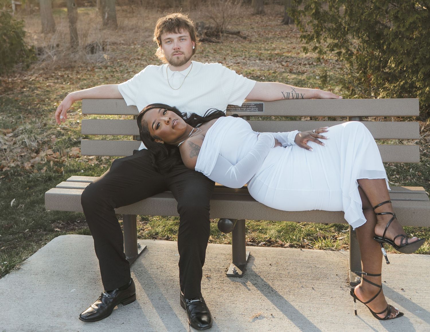 A man and woman pose on a park bench, both wearing white and black outfits, in a stylish outdoor photo shoot.