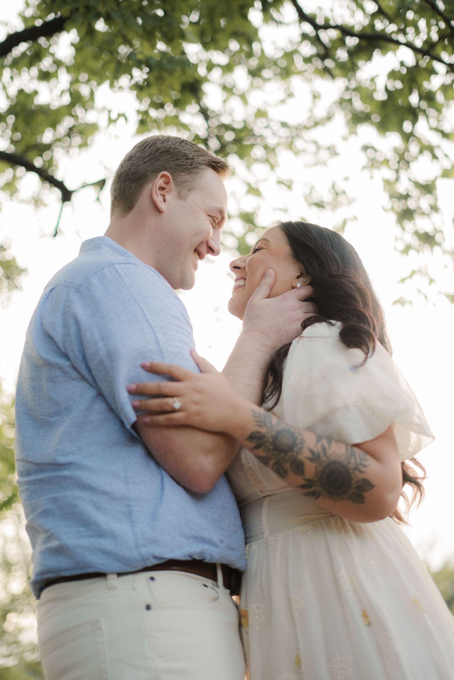 A couple shares a romantic moment outdoors, smiling at each other, the woman in a white dress with tattoos, the man in a blue shirt.