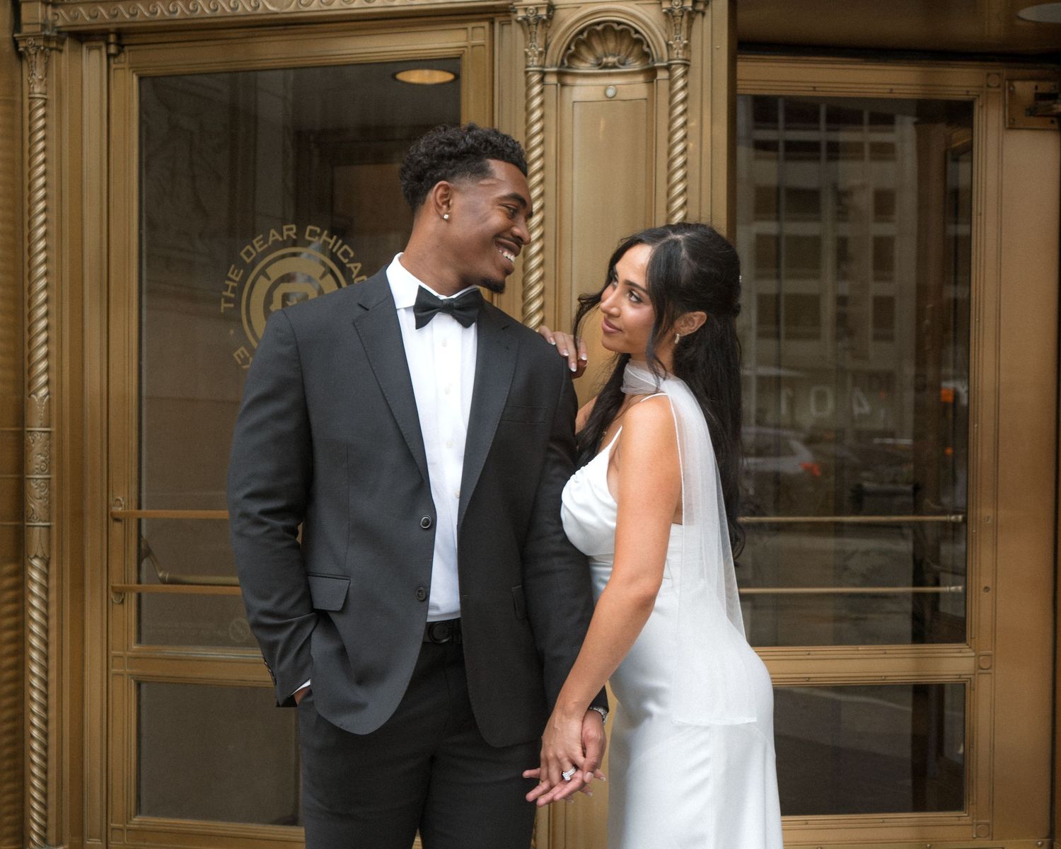 Elegant interracial newlywed couple holding hands and gazing at each other outside a gold revolving door.