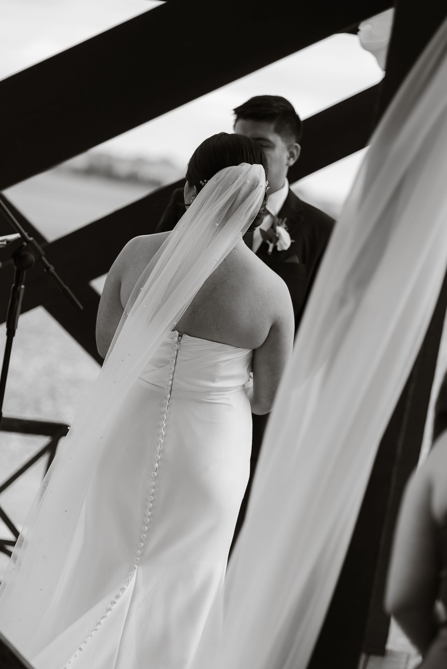 Black and white photo of bride and groom embracing, showcasing elegant backless wedding gown with veil and button details.