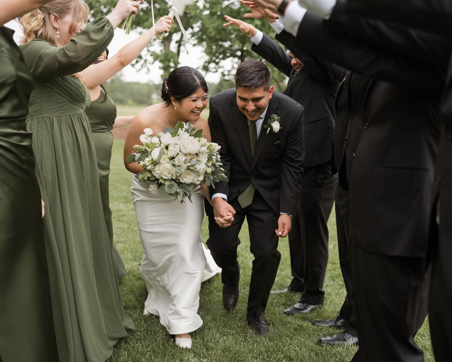 Bride and groom walk hand-in-hand through arch of raised streamers held by wedding party outdoors.