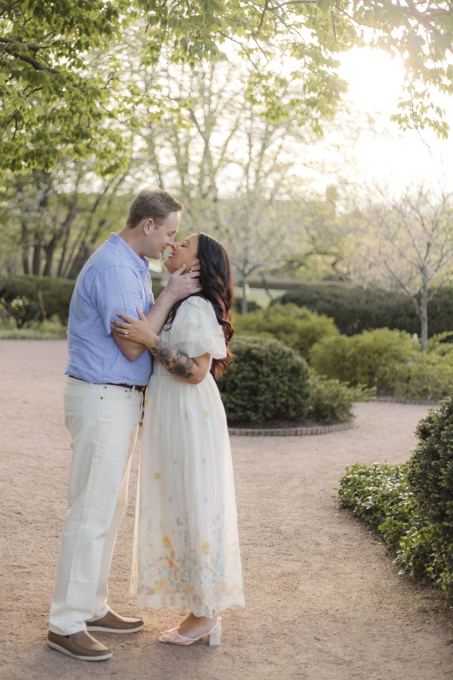 Couple sharing a romantic kiss in a sunlit garden, woman in floral dress, man in blue shirt, lush greenery backdrop.