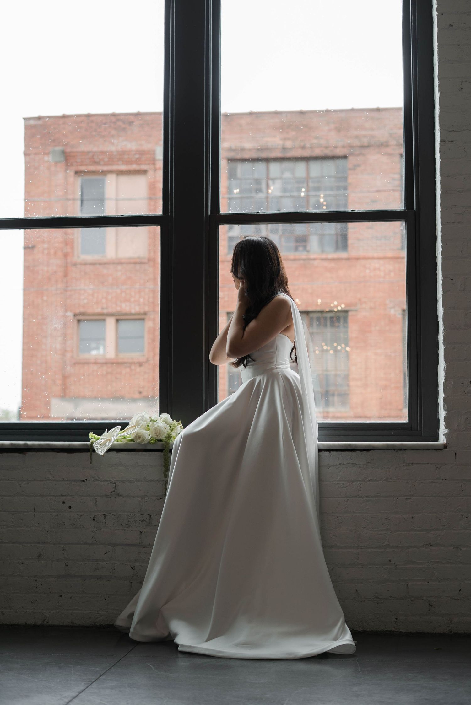 Bride in a sleek white wedding gown standing by large industrial windows holding white calla lilies bouquet.