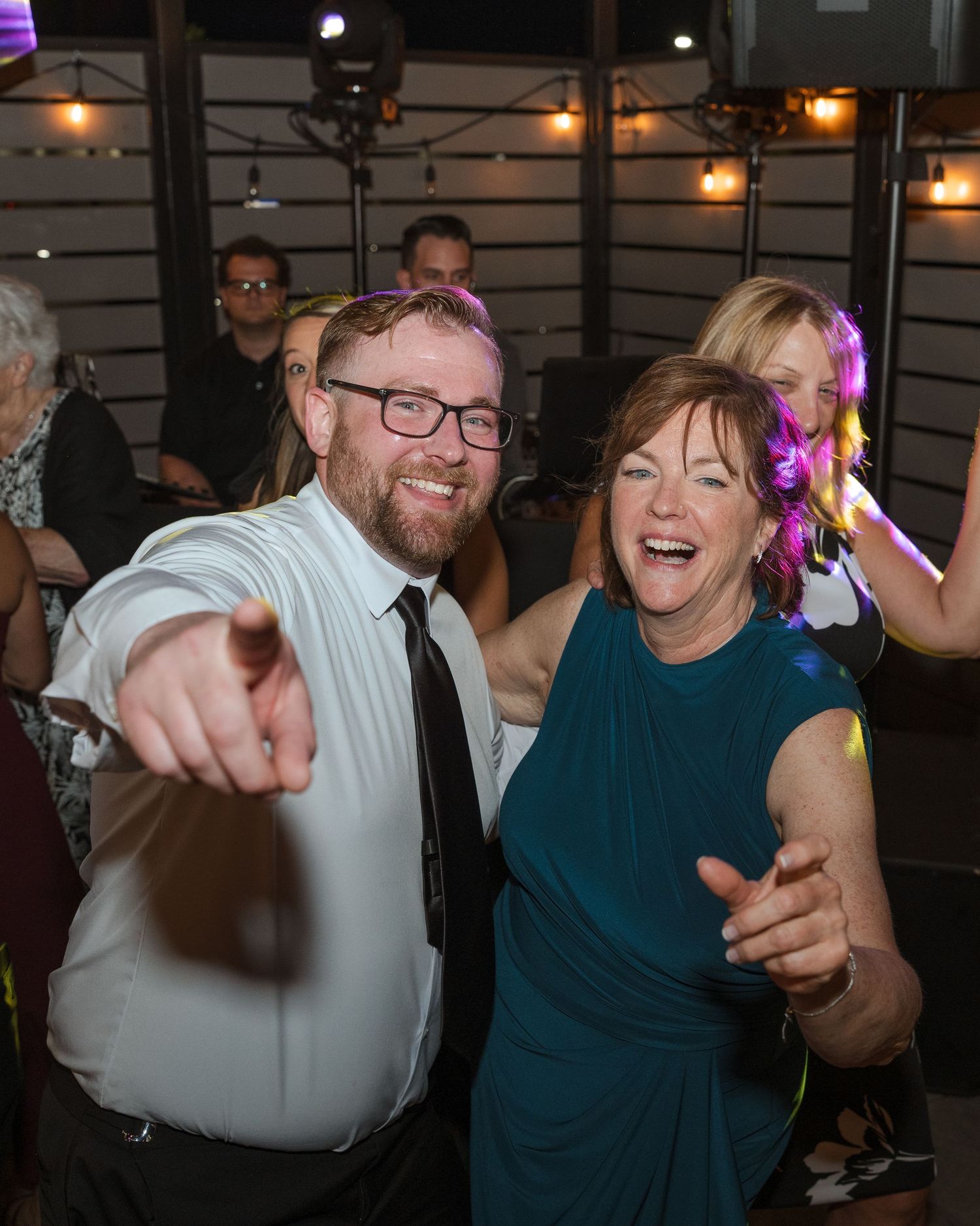 Two joyful people dancing and pointing at the camera at an indoor evening celebration or wedding reception.