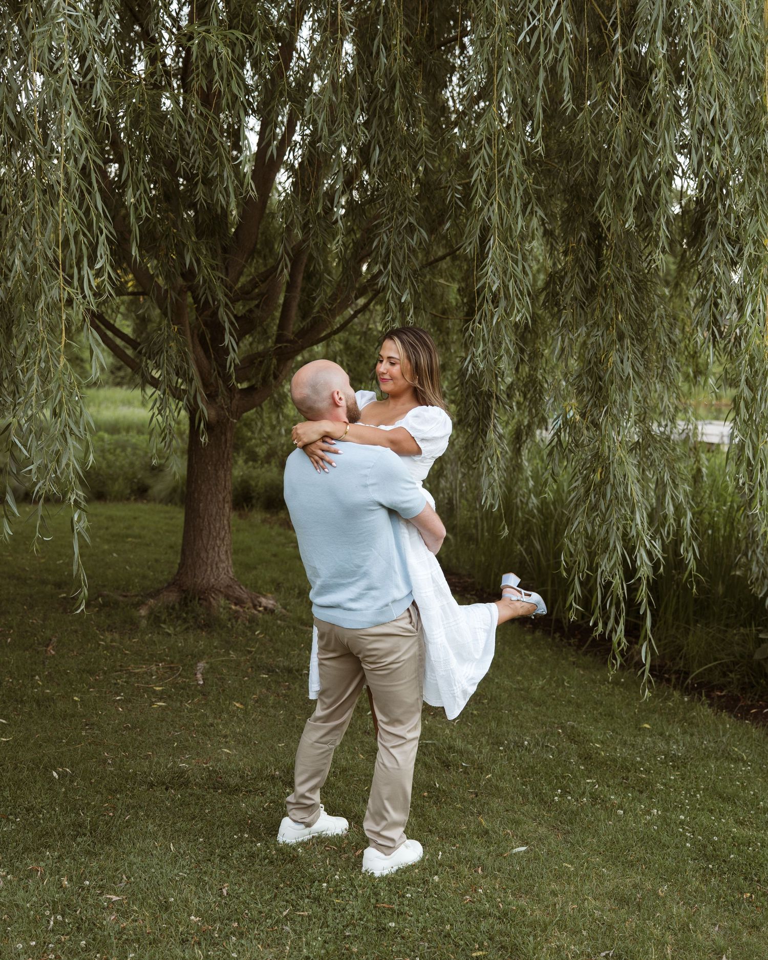 Man lifting woman in white dress outdoors near weeping willow tree during romantic engagement photo session.
