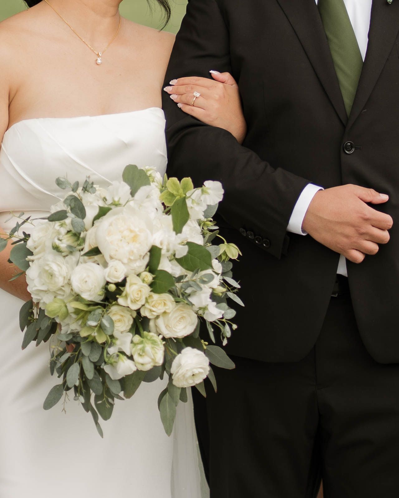 Bride in white strapless gown holding white floral bouquet stands beside groom in dark suit with green tie.
