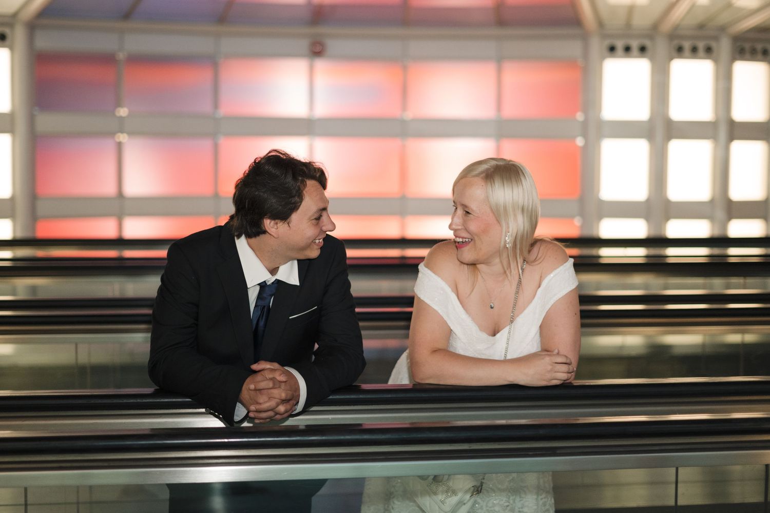 Couple in formal attire smiling at each other on an escalator with colorful red and orange background lighting.