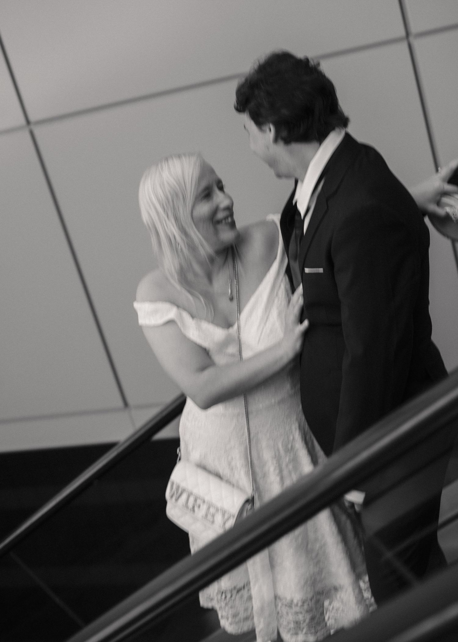 Black and white photo of a smiling blonde bride in white dress embracing a groom in suit on escalator.