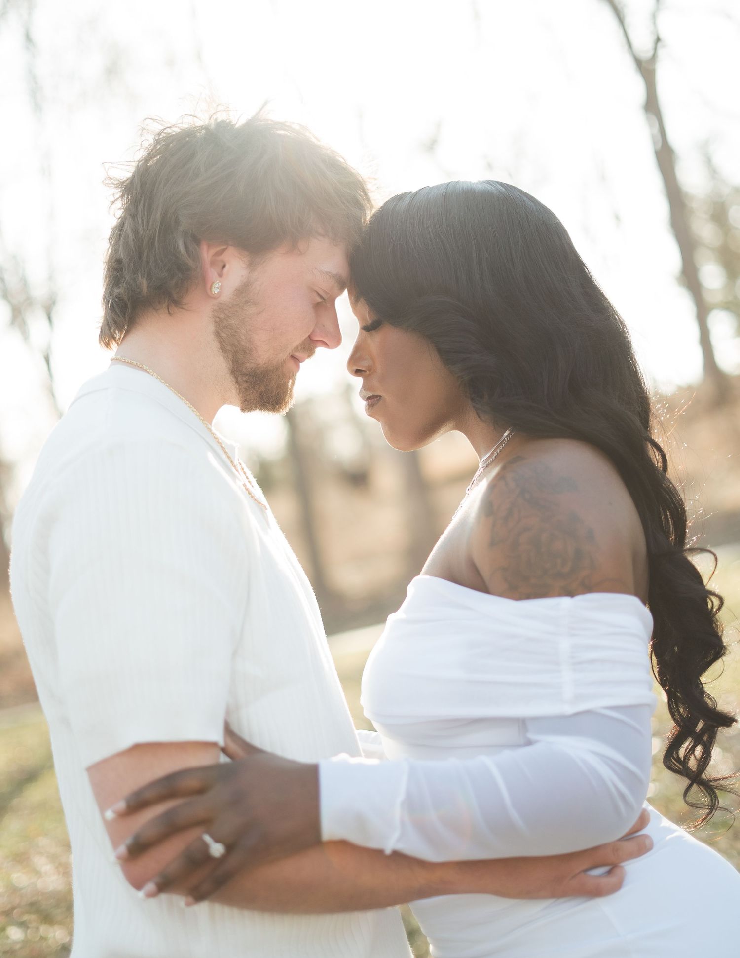 A romantic interracial couple in white outfits share an intimate moment outdoors in soft golden sunlight.