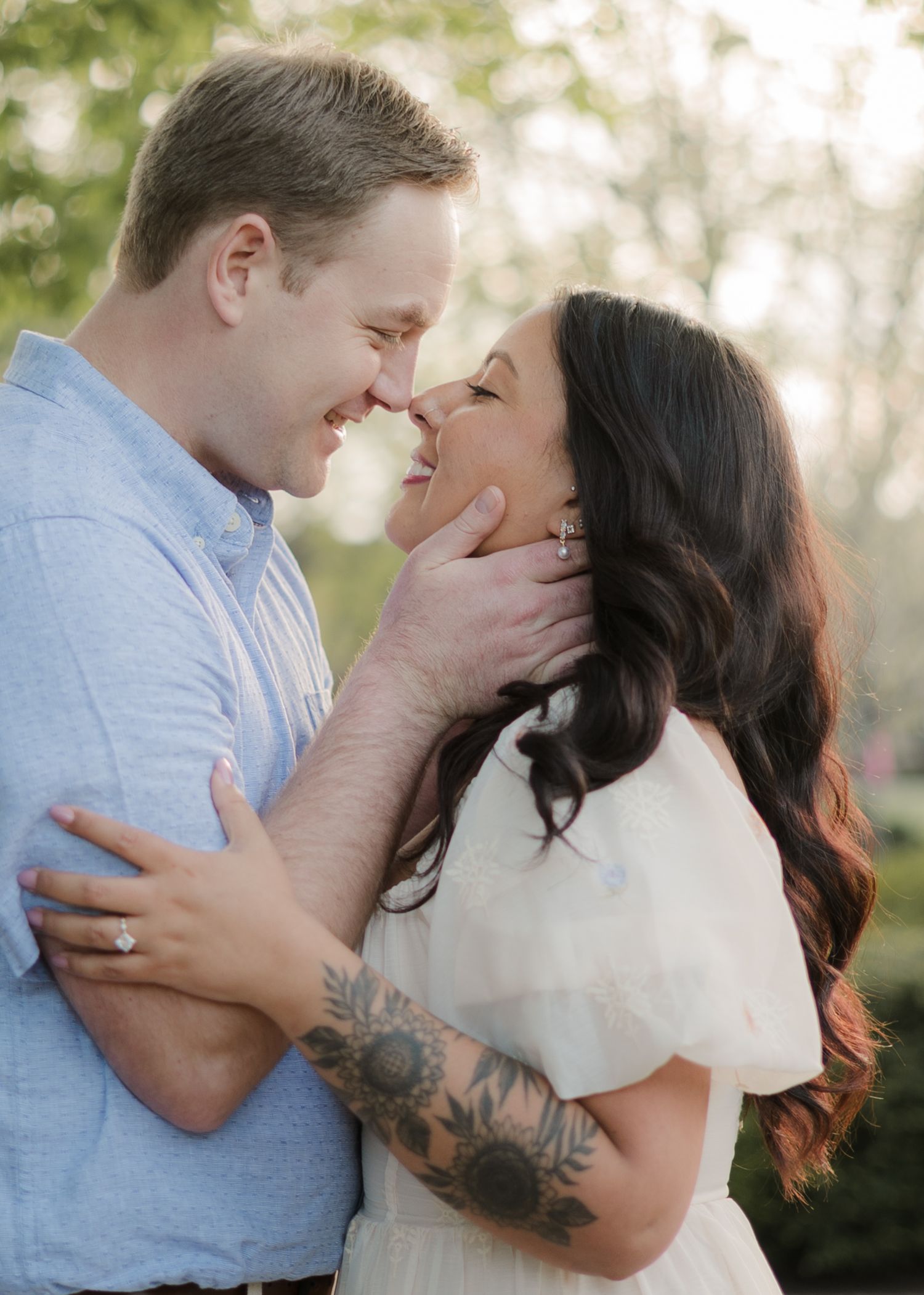 Couple sharing a romantic almost-kiss moment outdoors, woman with floral arm tattoo and engagement ring, soft natural lighting.