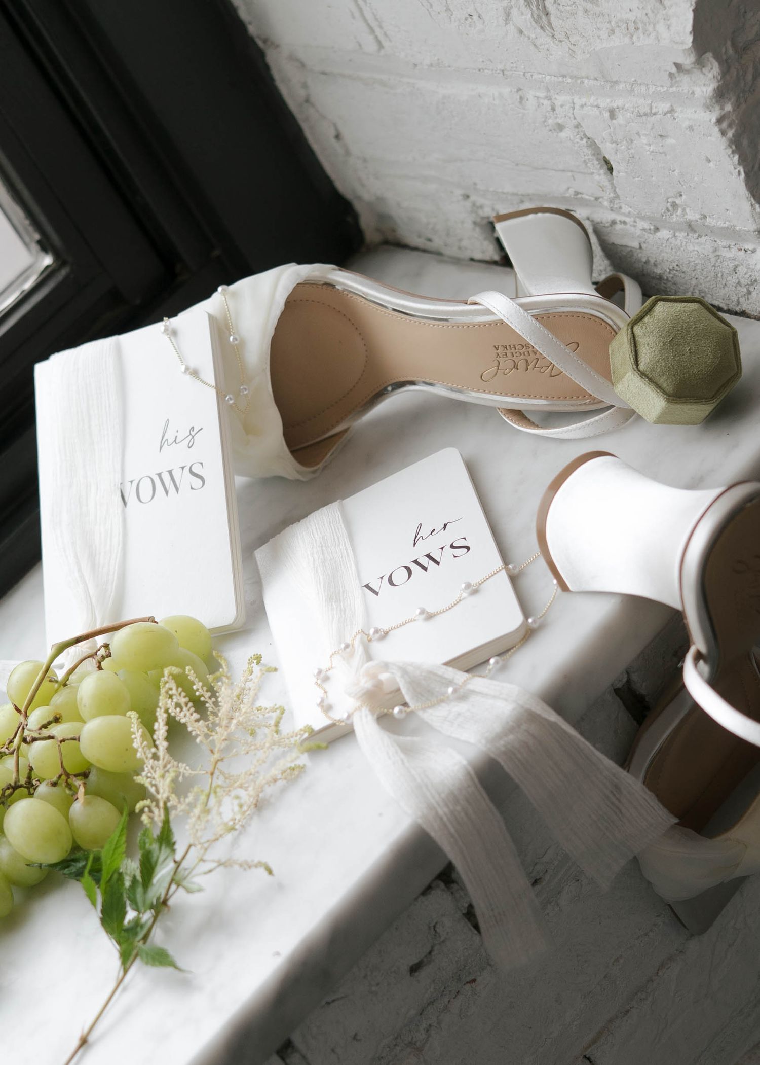 Elegant wedding flat lay with white heels, his and her vows books, green grapes, and floral sprigs on a white surface.