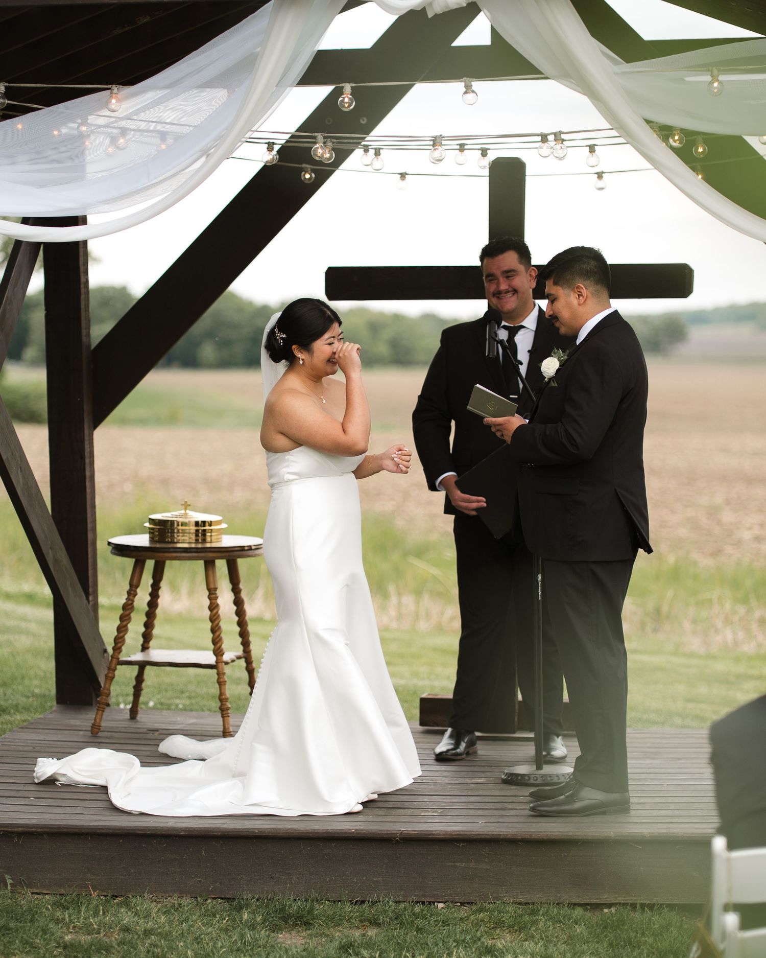 Bride and groom exchange vows under a rustic wooden gazebo with a cross during an outdoor wedding ceremony.