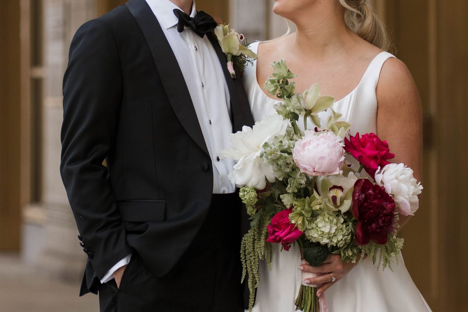 Bride and groom posing together, bride holding lush bouquet of pink, red, and white flowers with greenery on wedding day.
