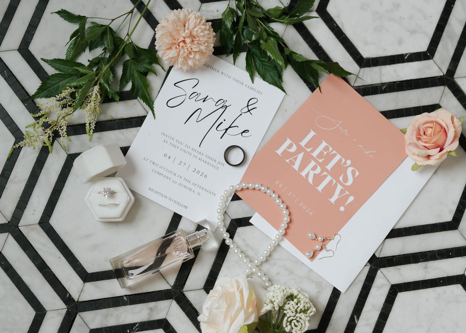 Wedding invitation suite with floral accents, ring box, pearl bracelet, and a pink party card on geometric marble.