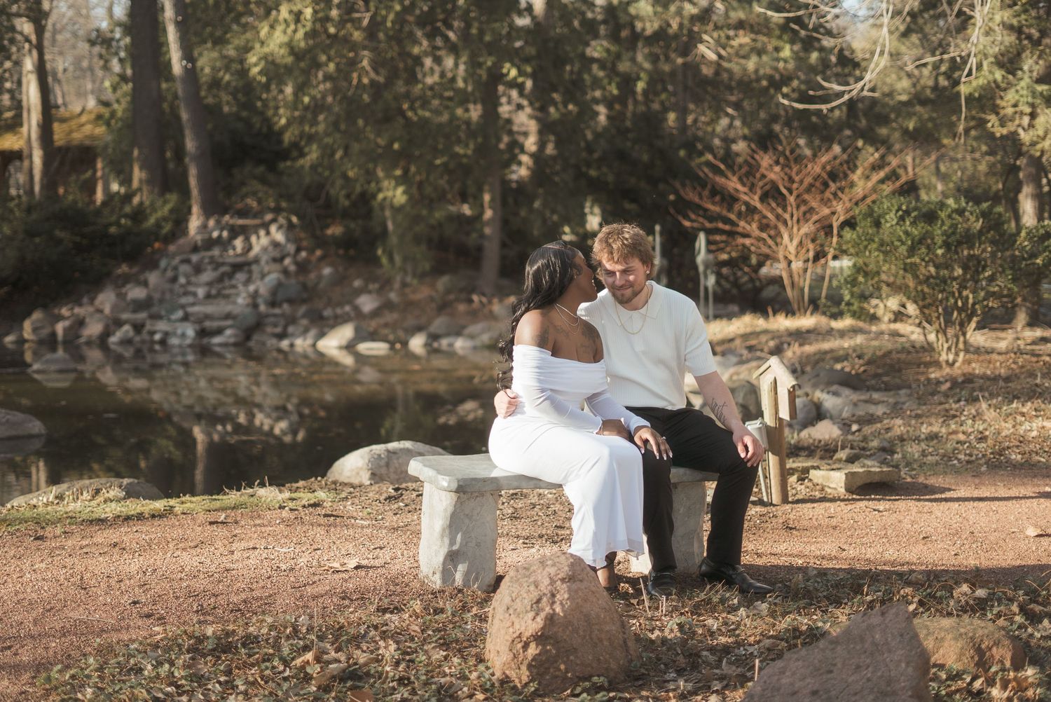 A couple sits closely on a stone bench in a serene park, sharing an intimate moment near a tranquil pond.