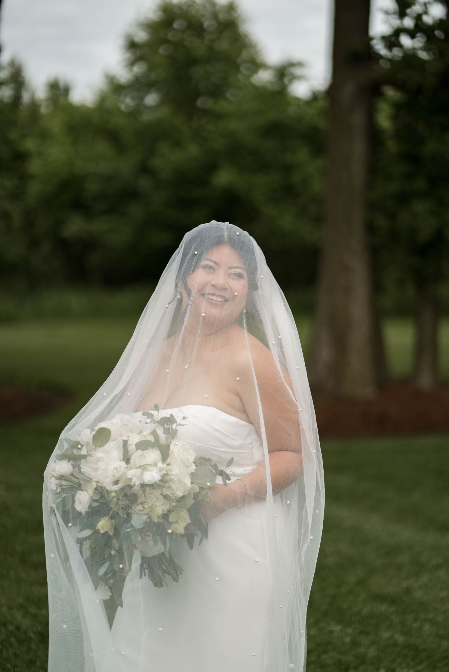 Smiling bride in white gown holding white bouquet draped under a pearl-dotted wedding veil outdoors.