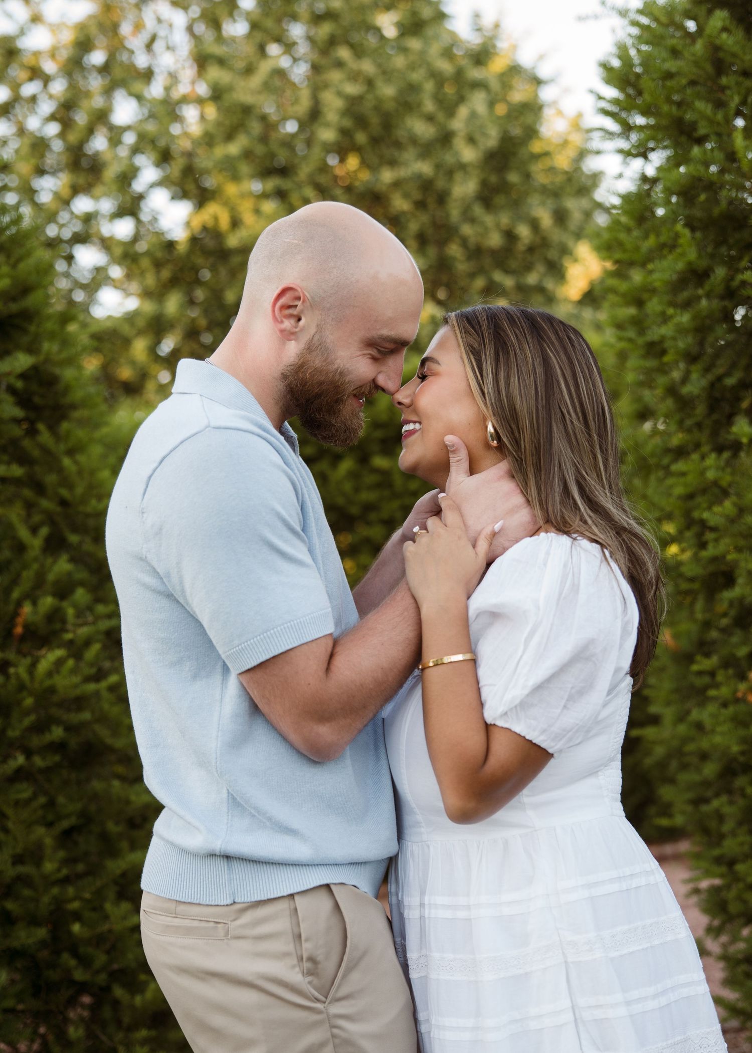 Smiling couple in light blue shirt and white dress sharing a tender moment among evergreen trees.
