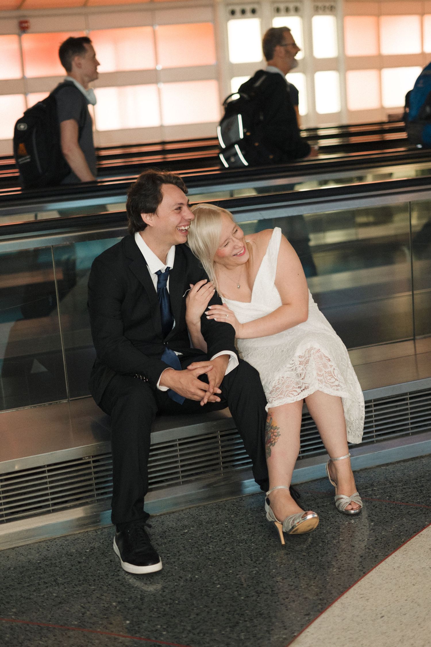 Happy couple in wedding attire sitting and laughing together on a moving walkway at an airport.