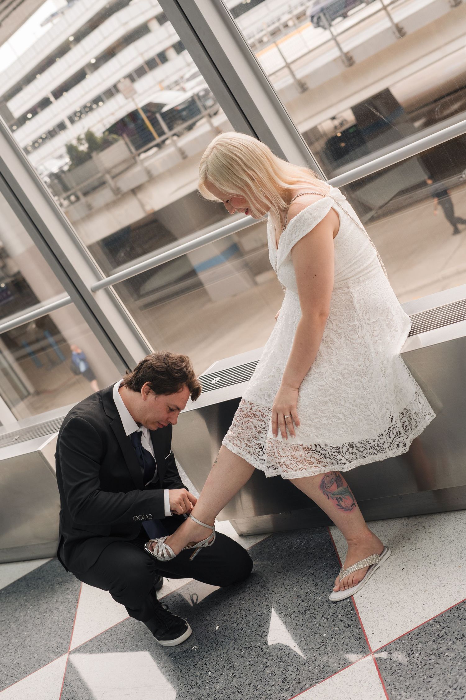 Groom kneeling to fix bride's shoe in white lace dress at modern venue.
