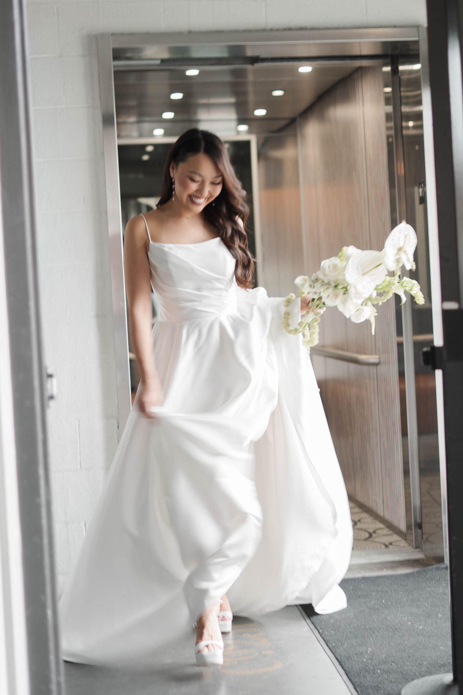 Happy bride in a flowing white spaghetti-strap wedding dress carrying an orchid bouquet through a glass door.