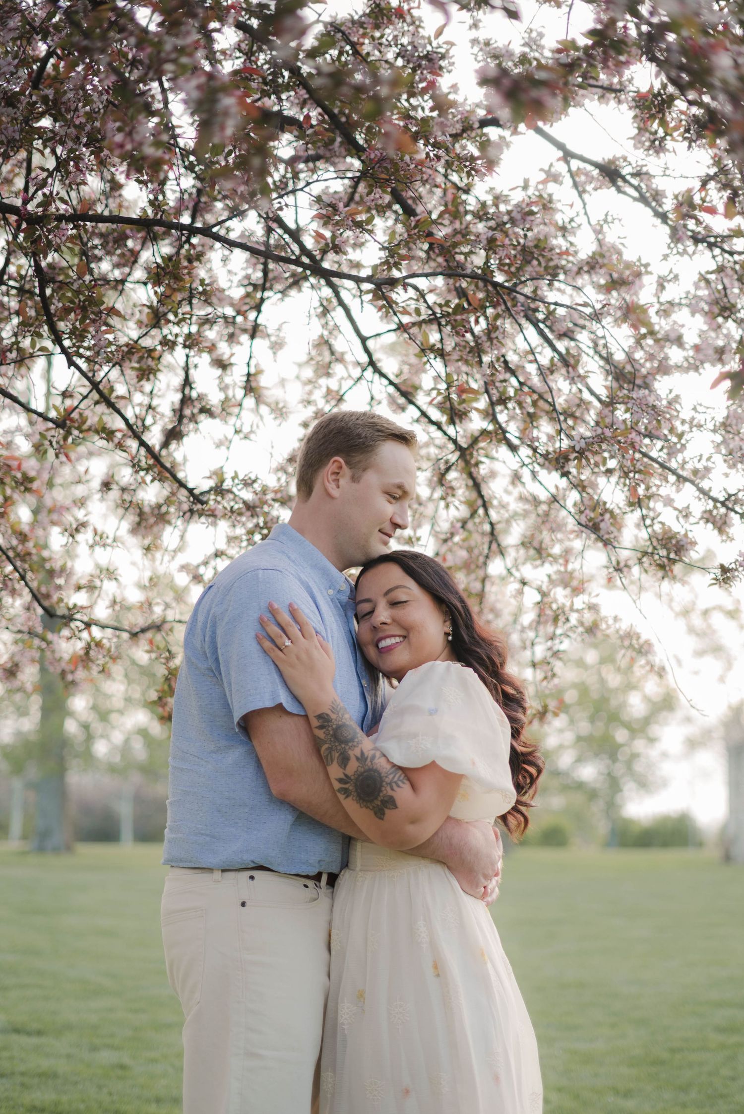 A smiling couple embraces under blooming cherry blossom trees in a park, wearing casual spring attire.