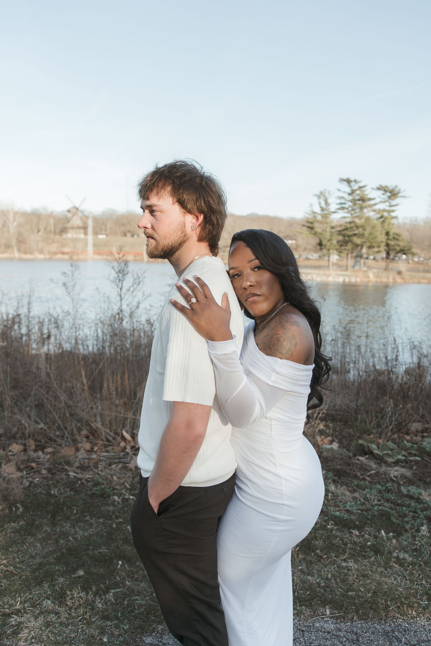 Interracial couple posing outdoors near a lake, woman in white dress embracing man from behind in a romantic maternity photo.