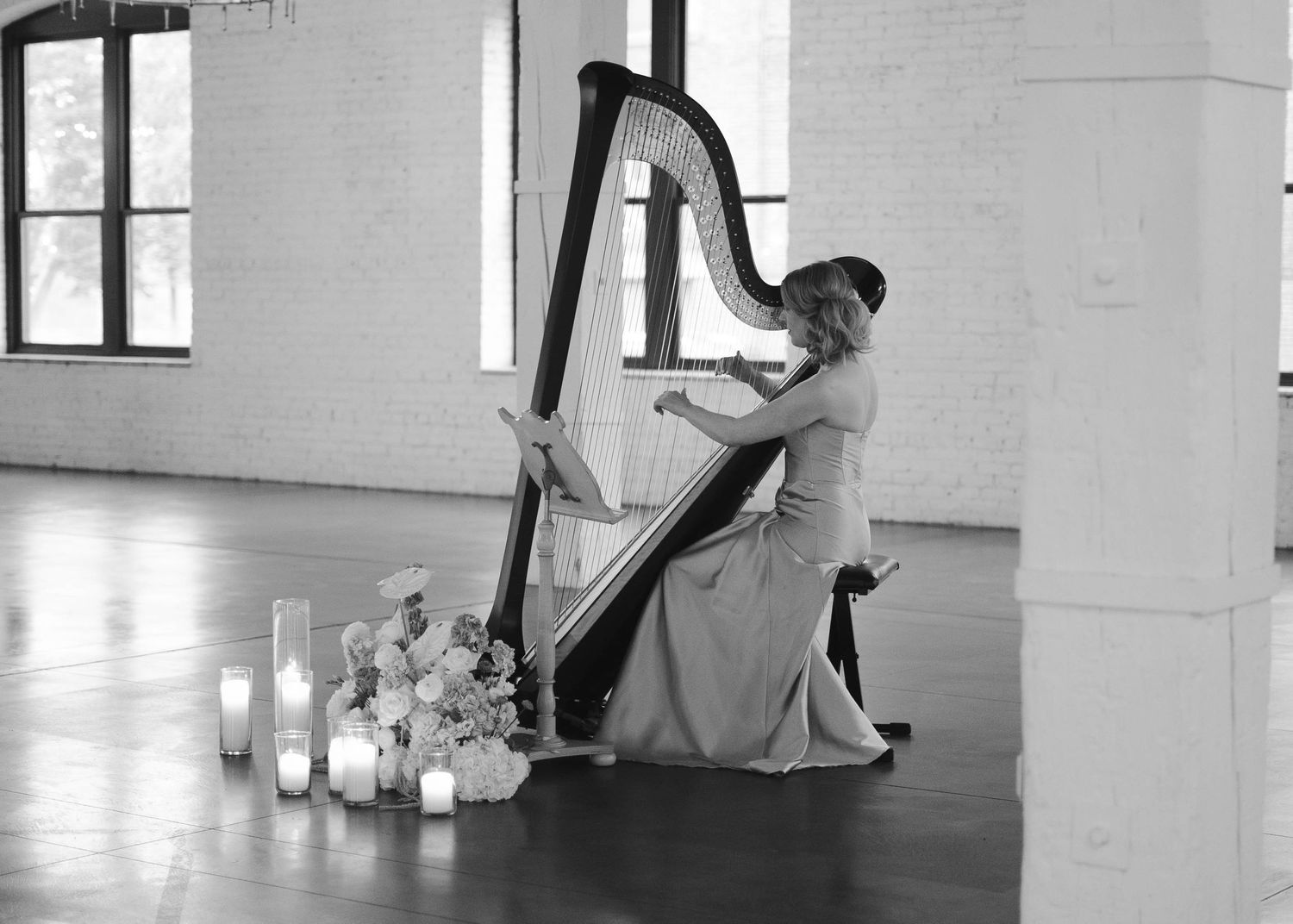 Elegant harpist performing at wedding ceremony in white brick venue surrounded by candles and flowers.