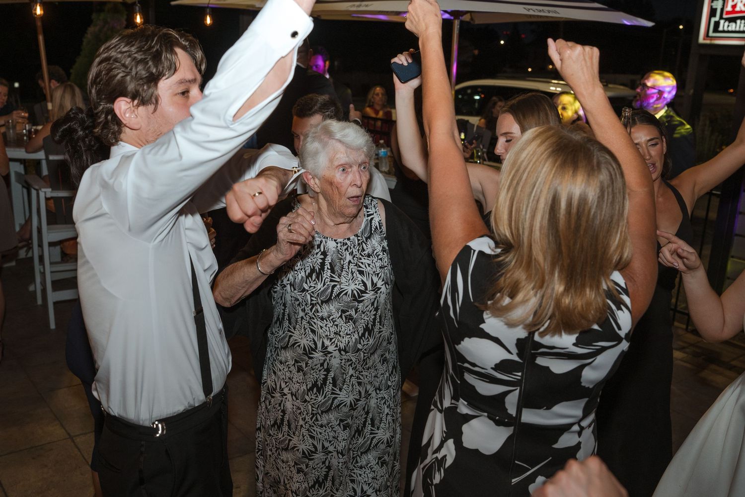 Elderly woman dancing joyfully with younger guests at an outdoor evening wedding reception party.